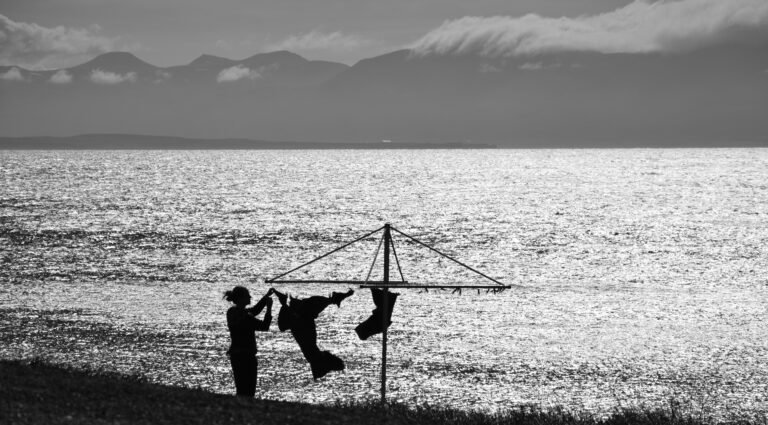 Dark silhouette of a woman hanging clothes to dry in front of a calm body of water.