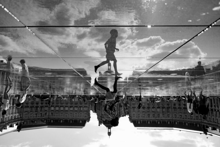 A boy appears upside down in a water reflection in front of a historic building in Bordeaux, photographed in black and white.